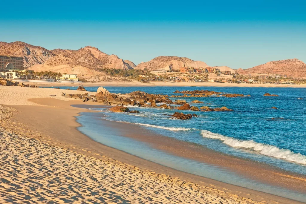 A serene beach in Cabo San Lucas with golden sand, gentle waves, and mountains in the background