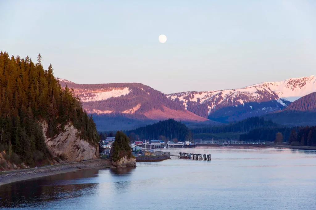 A serene dusk view of Icy Strait Point in Alaska, with a glowing moon above the calm sea