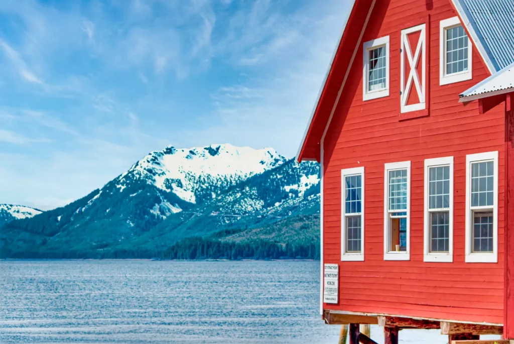 A vibrant red building overlooks a serene bay with snow-capped mountains in the background